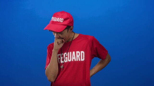Lifeguard man bites fist with hand on hip, wearing red cap and red shirt with lifeguard text and whistle around neck in studio; tired duty.