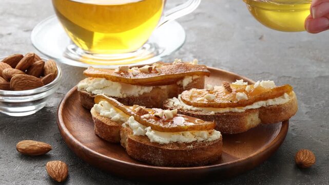 Woman dripping honey onto delicious bruschetta with cream cheese, pear and almonds at grey textured table, closeup