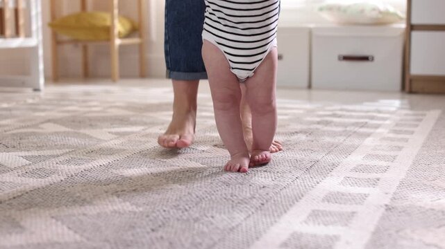First steps. Mother supporting daughter while she learning to walk at home, closeup