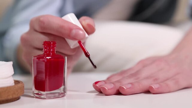 Woman applying red nail polish onto her nails at white table indoors, closeup