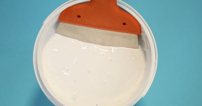 Bucket of plaster and putty knife on light blue background, top view. Camera spinning