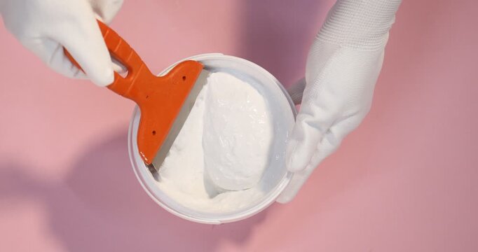 Worker with putty knife and bucket of plaster on pink background, top view
