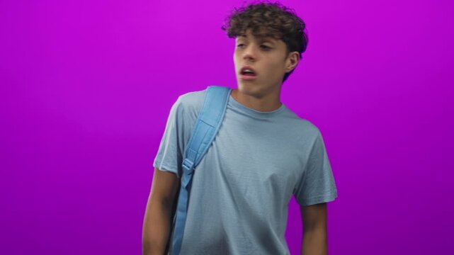Man with backpack looks to side in studio against purple backdrop, wearing blue tshirt and visible earring, head turned in profile and casual posture; curiosity.