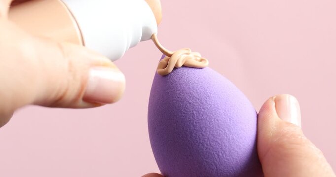 Woman applying skin foundation onto makeup sponge on pink background, closeup