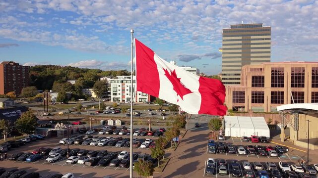 Canadian flag flutters strongly on Dartmouth shoreline with Halifax skyline and Atlantic Ocean in golden sunset light. Downtown buildings and bridge create scenic travel, culture and national identity
