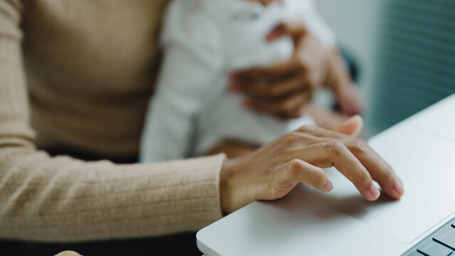 Mother holds child while typing on laptop. Soft focus highlights gentle care and work balance. Warm tones suggest comfort and calm focus. Perfect for parenting, remote work, or multitasking themes