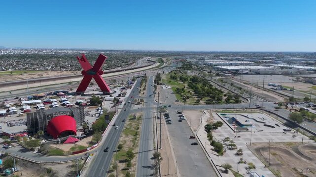 Aerial panoramic view of the red X monument at Plaza de la Mexicanidad, showing the border wall dividing Ciudad Juarez, Mexico, and El Paso, Texas, with heavy traffic on the highway.