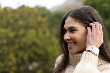 Naklejka premium Woman smiling tucking hair behind ear in park wearing cream turtleneck, pink smartwatch, copy space