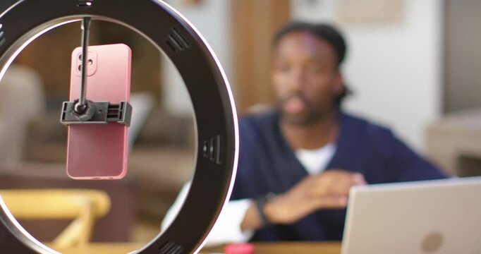 Man in scrubs at table showing bottle to camera as refocusing from ring light pink phone