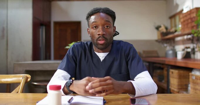 African American man in scrubs explaining medication, checking notes and gesturing at kitchen table