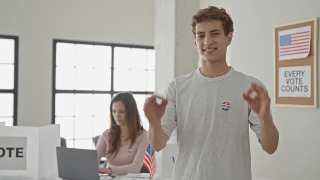Man wearing an i voted sticker makes ok gesture showing hands, standing near laptop and ballot box in a polling building; civic pride.