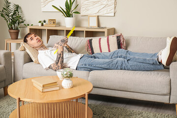 Young tattooed man with hand fan suffering from heat on sofa at home © Pixel-Shot