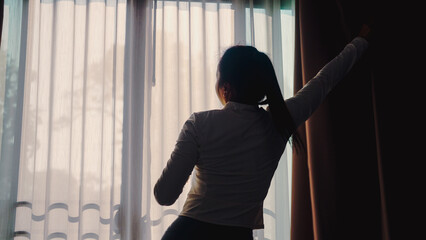 Woman reaches for curtain, sunlight streams through sheer drapes. She stands by window, back to camera, arms extended. Soft light illuminates her silhouette, quiet moment. Ideal for home, morning © WARAPHON
