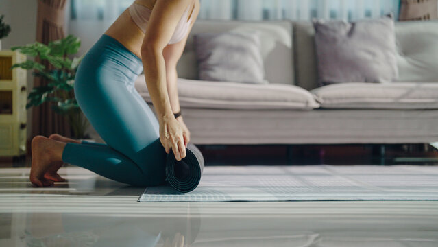 Woman kneels on floor, rolling yoga mat. Soft light fills cozy living room space. She focuses, preparing for gentle stretching. Mat rests on polished, reflective surface