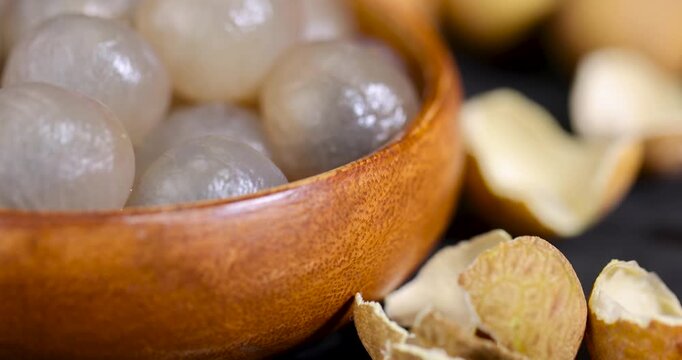 A light peeled longan in a bowl, a full bowl of longan fruit kernels stands on the black surface of the slate near unpeeled whole fruits.