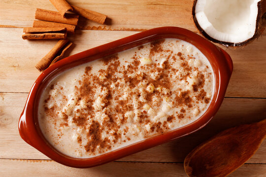 A bowl of creamy white corn porridge with cinnamon, accompanied by peanut brittle and coconut, served on a wooden base. A popular traditional Brazilian