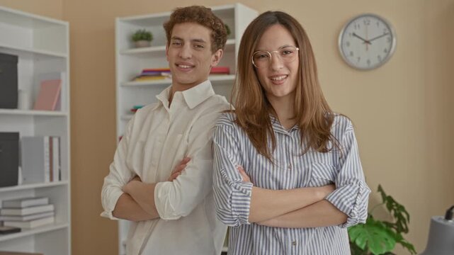 Man and woman stand shoulder to shoulder with arms crossed, smiling, beside a bookshelf and wall clock in a building workspace; teamwork confidence success.
