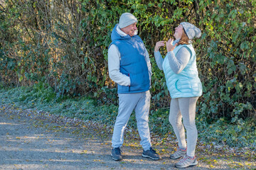 Fototapeta premium two adults in warm clothes talking outdoors near green bushes, man smiling while woman gestures energetically during walk, outdoor communication, active conversation, friendly communication moment