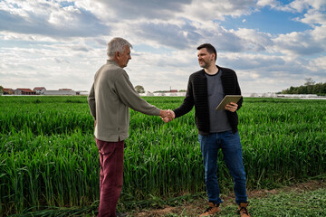 Two men shaking hands on agricultural field, younger man holding tablet. Expert and farmer or selling property concept © phoenix021