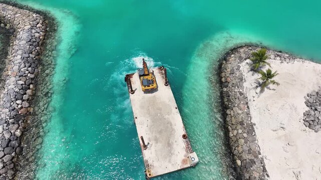 Aerial view of an excavator on a barge working in turquoise waters between stone breakwaters near a white sand beach in Kamadhoo, Baa Atoll, Maldives.