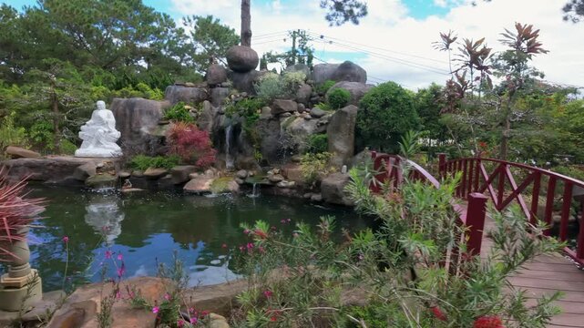 A picturesque red bridge arches over a tranquil pond with a small waterfall in a lush garden in Thien Vien Truc Lam, Da Lat, Vietnam. The serene landscape invites peaceful contemplation.