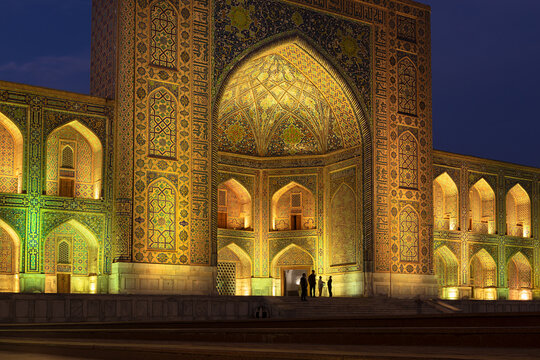 Illuminated facade of Tilla-Kari Madrasah at Registan Square in Samarkand at twilight. Intricate tilework, pointed arches glow under warm lighting against a deep blue sky. Ideal for travel, heritage.