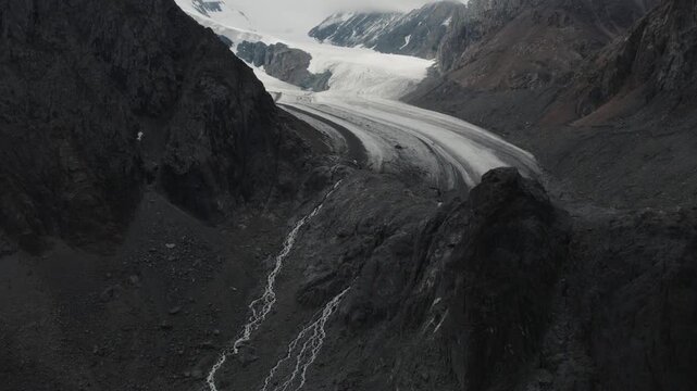 Drone footage of a glacier tongue flowing through the Aktru Valley, surrounded by dark rocky peaks and meltwater streams in the Altai Mountains, Altai Republic, Russia