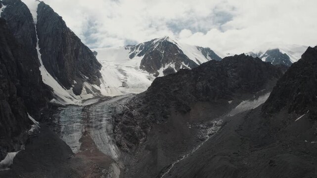 Drone footage of the Aktru Valley with a large glacier, dark rocky ridges and snowfields under cloudy sky, Altai Republic, Russia, remote alpine wilderness