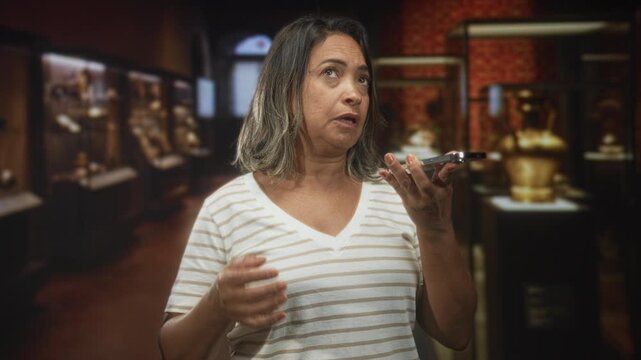 Middle age hispanic woman holds a smartphone in her hand while speaking into it with a raised hand and looking up inside a museum building; curiosity.