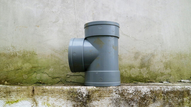 Gray pvc sewer pipe tee fitting on weathered concrete ledge against stained plaster wall industrial plumbing component
