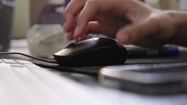 a hand using a wired computer mouse on a desk in a bright office. Features a white keyboard, shallow depth of field, and a professional workspace setting for technology themes.
