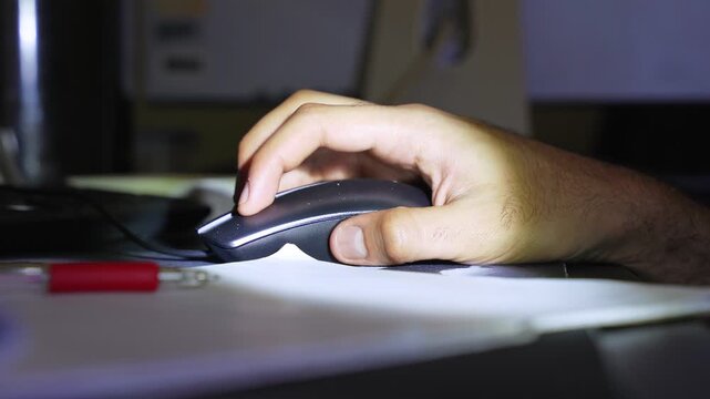 a hand using a black computer mouse on a desk at night. Low-light cinematic scene with a red pen in the blurred foreground, ideal for remote work or late-night study themes.