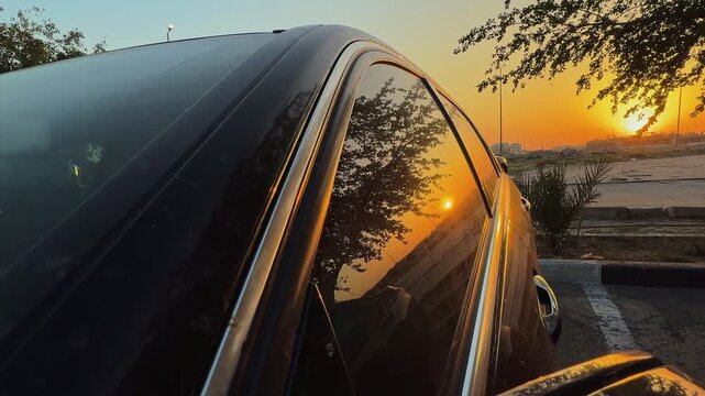 a golden sunset reflecting on a sleek black car window in an outdoor setting. Warm orange light glows on the luxury vehicle body during golden hour in a cinematic atmosphere.