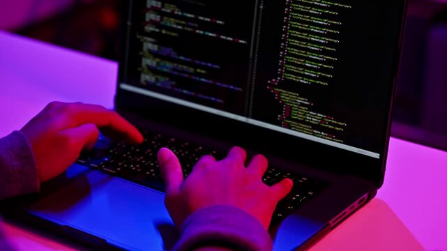 Close up of hands typing on laptop keyboard with programming code on screen in dark neon lit room
