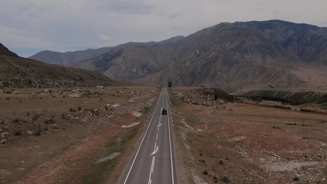 Drone footage of a car driving along Chuysky Trakt, a scenic mountain highway across a wide valley with rocky hills and river landscape in Altai Republic, Russia
