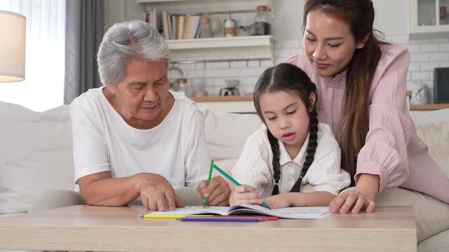 Happy Asian three generation family drawing together in living room, Smiling grandmother, mother and little daughter coloring book on table, Warm home education, bonding and family leisure concept