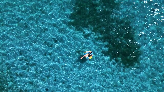 Aerial view of a woman floating in a blue and yellow inflatable ring on crystal clear turquoise water with sunlight ripples Guanaja, Bay Islands Department, Honduras.