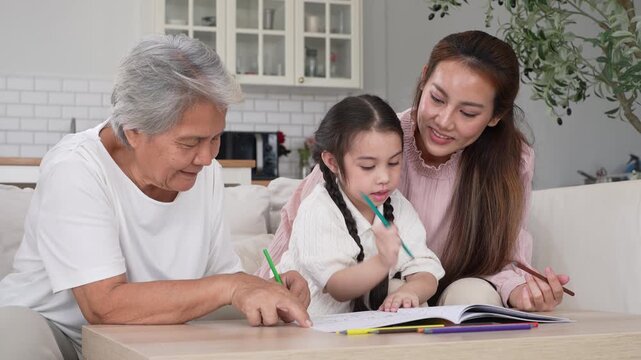 Happy Asian three generation family drawing together in living room, Smiling grandmother, mother and little daughter coloring book on table, Warm home education, bonding and family leisure concept