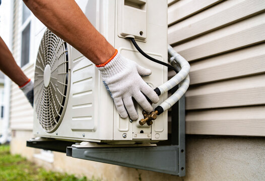A person's gloved hands are working on an outdoor air conditioning unit, connecting pipes to a house wall.