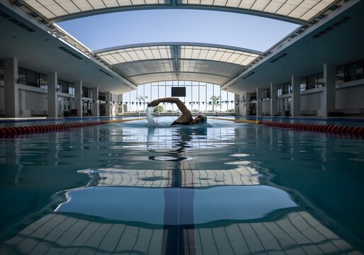 Man swimming in a modern indoor swimming pool, sunlight reflects off the water surface.