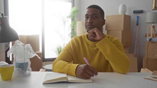 Black man writing in notebook with pen, hand to chin gesture in building among moving boxes, lamp and bubblewrap; thoughtful planning.