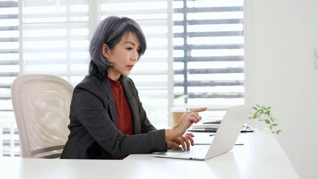 Mature senior asian professional business woman working on laptop, looking at screen searching information at office.