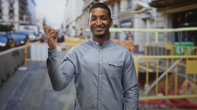 Man pointing finger toward yellow construction barrier on urban street while smiling and looking at camera; direction guidance confidence.