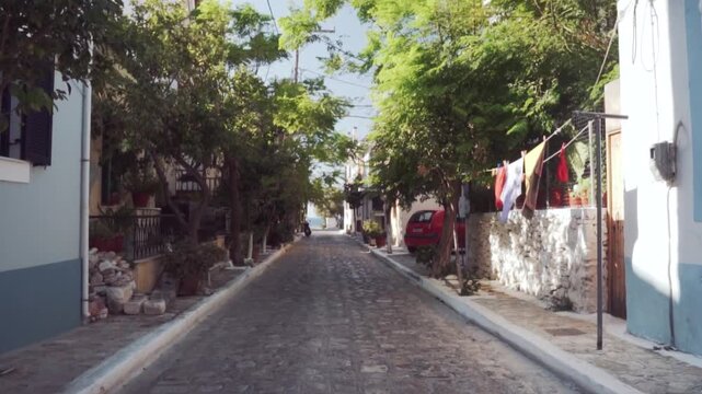 Ground-level view of a sunlit cobblestone street in Molyvos, Lesvos, lined with whitewashed houses, vibrant green trees, potted plants, and laundry gently swaying, with no people present. (200)