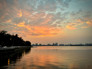 Naklejka premium Peaceful Lakeside Sunset With Orange Clouds, Calm Water, and Distant Urban Skyline