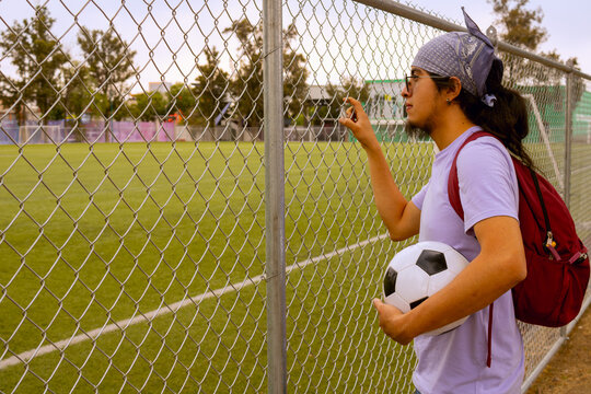 Young Soccer Player with Backpack Looking at Football Field Through Fence