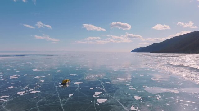 Aerial view of a hovercraft traveling across clear cracked ice on Lake Baikal in Siberia, Russia, with snowy mountains, blue sky and scattered clouds
