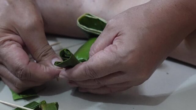Hands folding and pinning a green banana leaf with a wooden stick, finished with a scissor cut.
