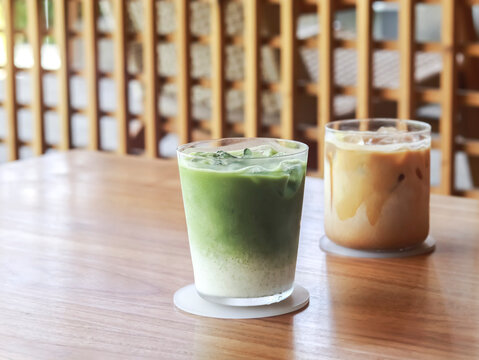 Iced matcha green tea latte and an iced caramel coffee latte on a warm wooden table in a cafe, with blurred light-colored wood lattice in the background.