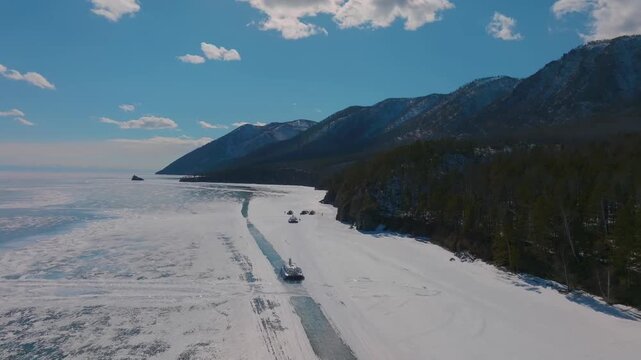 Aerial view of hovercraft and vehicles traveling on an ice road across frozen Lake Baikal, Siberia, Russia, with snowy mountains and clear blue sky in winter